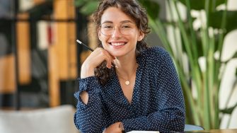 Portrait of smiling woman wearing spectacles and holding pencil while sitting at desk. Business woman taking notes in diary and looking at camera. University girl with eyeglasses sitting on table at library.