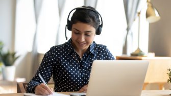 Happy indian girl student wear headset study online with webcam teacher write notes, happy young woman listen lecture watch webinar on laptop sit at desk, distance e learn language education concept