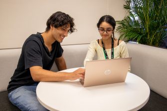 Two students sitting next to each other, both smiling and looking at a laptop screen.