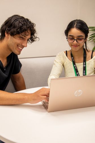 Two students sitting next to each other, both smiling and looking at a laptop screen.