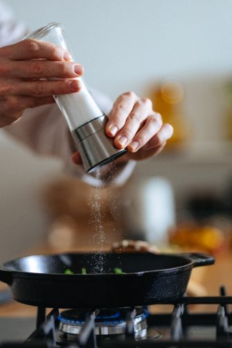 An anonymous chef with apron standing at the gas stove and pouring salt into frypan.