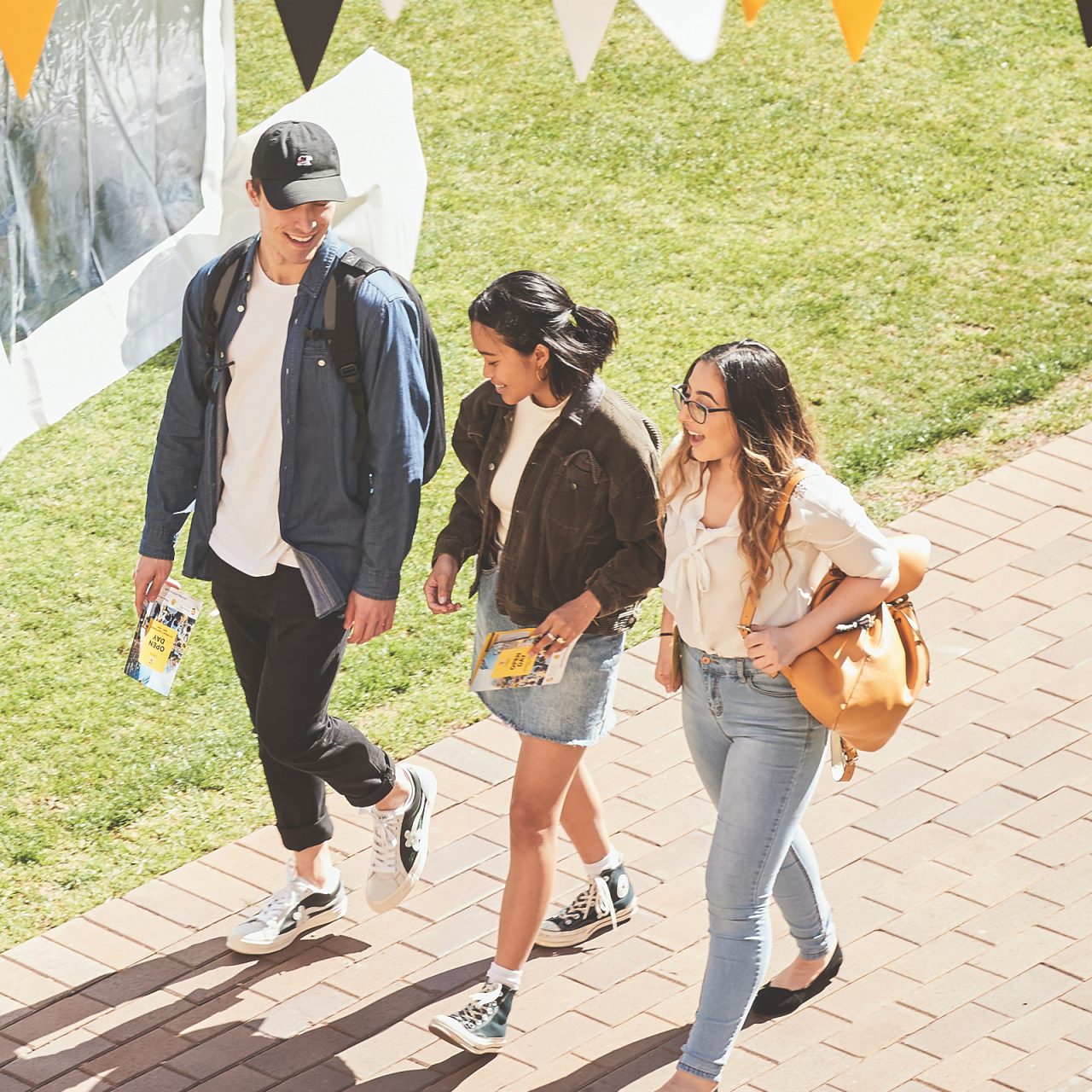 Three happy students walk along the pathway through Kensington Quad on a sunny day
