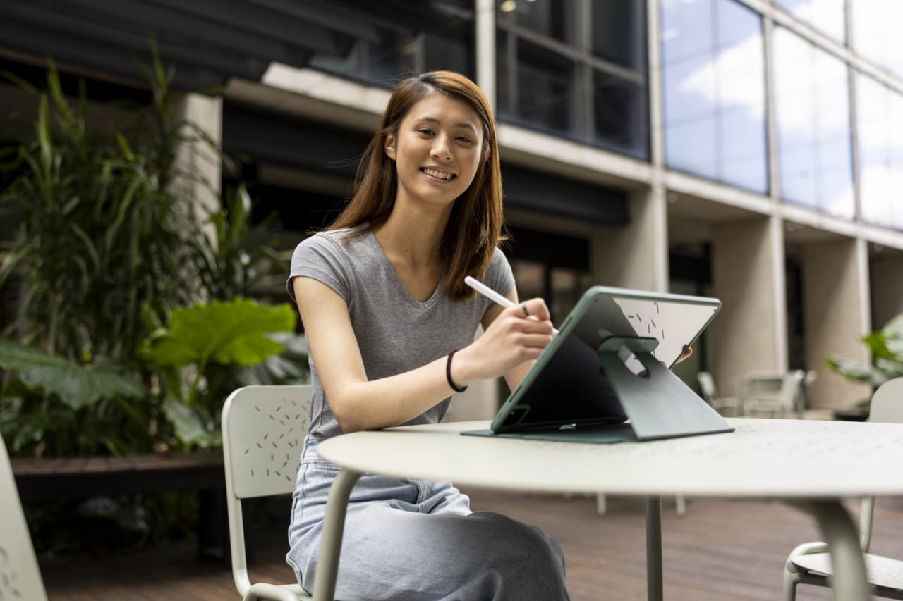 Smiling student using iPad with pen in courtyard